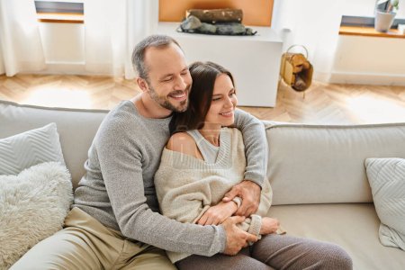 hombre feliz con los ojos cerrados abrazando esposa sonriente de sofá acogedor en la sala de estar, estilo de vida libre de niños