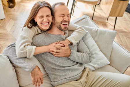 mujer excitada con los ojos cerrados abrazando marido riendo en el sofá en la sala de estar, pareja libre de niños