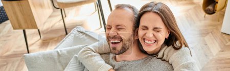 mujer alegre con los ojos cerrados abrazando al hombre riendo en el sofá en casa, pareja sin niños, pancarta