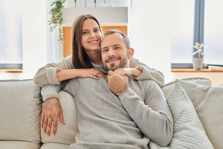 happy woman hugging smiling husband sitting on couch and looking at camera, child-free lifestyle