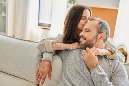 glückliche Ehefrau küsst den Kopf eines lächelnden Ehemannes, der mit geschlossenen Augen auf der Couch sitzt, kinderfreier Lebensstil