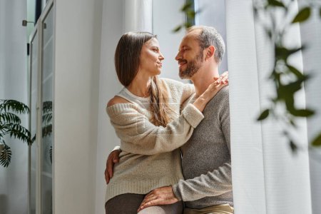joyful child-free couple hugging and looking at each other near window in cozy living room, warmness
