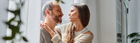 happy child-free couple hugging and looking at each other near window in cozy living room, banner