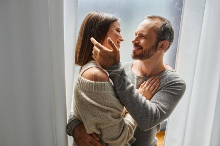 loving child-free couple embracing and smiling at each other near window at home, tenderness