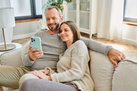 Lächelnder Mann surft im Internet auf Smartphone neben Frau auf Couch im Wohnzimmer, kinderfreies Paar