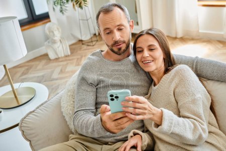 joyful child-free couple relaxing on couch at home and browsing internet on smartphone, leisure