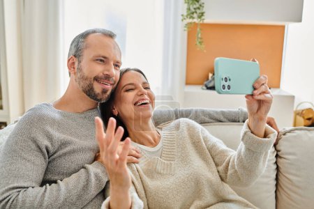 cheerful woman taking selfie on smartphone with husband in living room, leisure of child-free couple