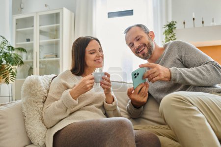 excited man showing smartphone to wife with coffee cup on couch in living room, child-free couple