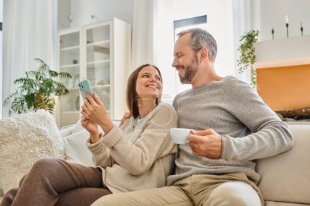 glückliches kinderfreies Paar mit Kaffeetasse und Smartphone, das sich auf der Couch im Wohnzimmer anschaut