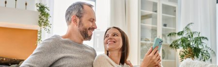 pleased looking at pleased wife with smartphone at home, child-free lifestyle, horizontal banner