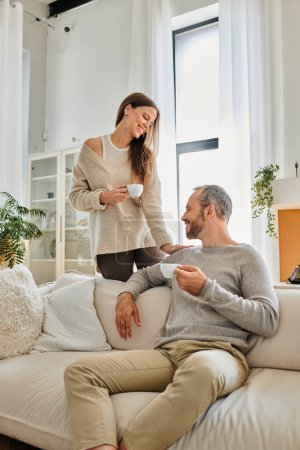joyous child-free couple drinking morning coffee in modern living room of cozy apartment, serenity