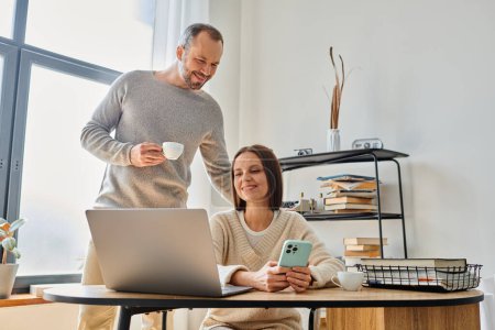 Lächelnder Mann mit Kaffeetasse unterstützt Ehefrau bei der Arbeit am Laptop zu Hause, Einheit des kinderlosen Paares