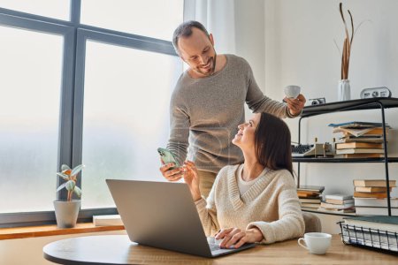 fröhlicher Mann mit Kaffeetasse zeigt Smartphone seiner Frau, die zu Hause am Laptop arbeitet, kinderfreies Paar
