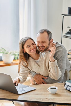 aufgeregter Mann umarmt glückliche Ehefrau, die zu Hause am Laptop arbeitet, Pflege und Unterstützung kinderloser Paare