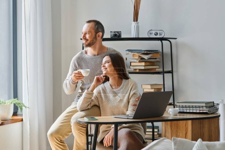 cheerful man with coffee cup and woman at laptop looking away in office, happy child-free couple