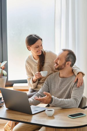 glückliche Frau mit Kaffeetasse unterstützt Ehemann bei der Arbeit am Laptop im Homeoffice, kinderfreies Paar