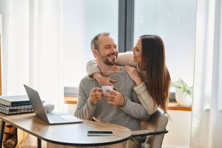 fröhlicher Mann mit Kaffeetasse neben Laptop sitzend und fürsorgliche Frau im Home Office, kinderfreies Paar