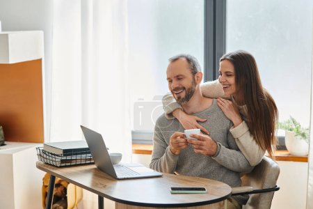 freudige Frau umarmt lächelnden Ehemann sitzt mit Kaffeetasse neben Laptop, kinderfreier Lebensstil