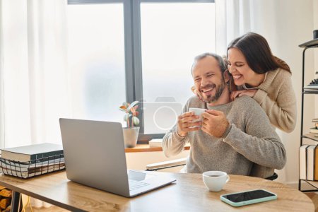 fröhliche Ehefrau umarmt lächelnden Ehemann sitzt mit Kaffeetasse neben Laptop, kinderfreier Lebensstil