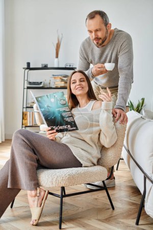 Téléchargez les photos : Femme heureuse lisant le magazine scientifique près du mari avec tasse de café sur le canapé à la maison, vie sans enfant - en image libre de droit