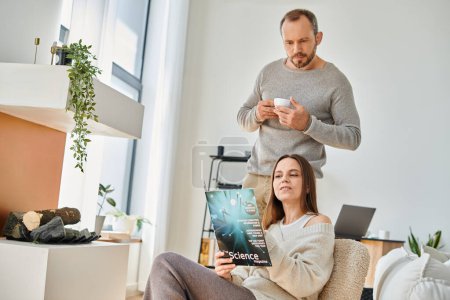 interessierte Frau liest Wissenschaftsmagazin in der Nähe Ehemann mit Kaffeetasse auf Couch, kinderfreies Paar