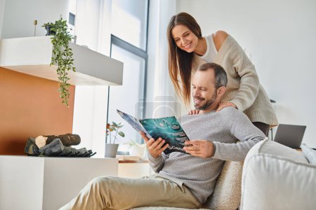 freudiger Mann liest Wissenschaftsmagazin neben lächelnder Frau auf Couch im Wohnzimmer, kinderfreies Paar