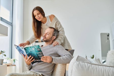 glücklicher Ehemann liest Wissenschaftsmagazin neben lächelnder Frau auf Couch im Wohnzimmer, kinderfreies Paar
