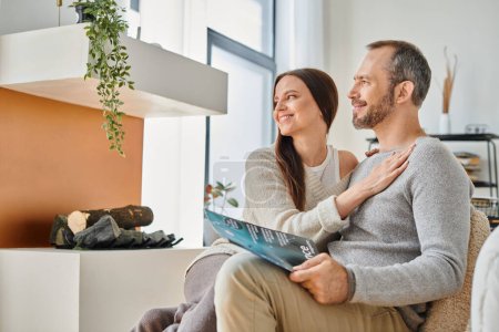 liebevolle Ehefrau umarmt Ehemann sitzt mit Wissenschaftsmagazin auf Couch zu Hause, kinderfreies Leben