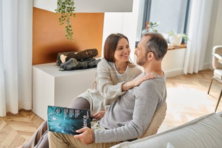 fröhliche Frau umarmt Ehemann sitzt mit Wissenschaftsmagazin auf Couch zu Hause, kinderfreies Paar