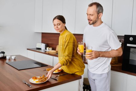 Téléchargez les photos : Homme avec du jus d'orange près d'une femme souriante servant un délicieux petit déjeuner dans la cuisine, couple sans enfant - en image libre de droit