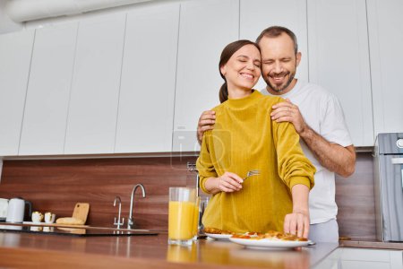joyful man with closed eyes hugging shoulders of wife near breakfast in kitchen, child-free couple