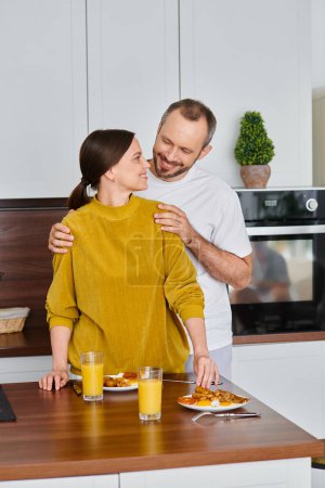 lächelnder Mann umarmt Schultern seiner Frau, die das Frühstück in der Küche serviert, am Morgen des kinderfreien Paares