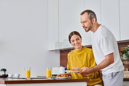 mari servant un délicieux petit déjeuner près de femme heureuse dans la cuisine, matin de couple sans enfant