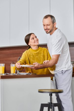 Téléchargez les photos : Femme étonnante souriant près du mari servant un délicieux petit déjeuner dans la cuisine, couple sans enfant - en image libre de droit