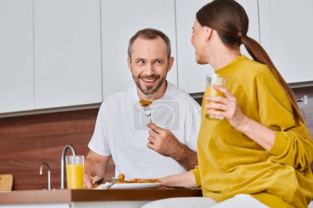 fröhlicher Mann mit leckerem Frühstück in der Nähe seiner Frau mit Orangensaft in der Küche, kinderfreier Lebensstil