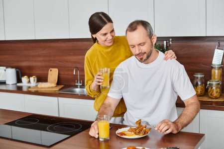 caring wife holding orange juice near pleased husband enjoying breakfast in kitchen, love and care