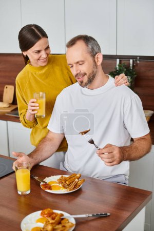 happy wife holding orange juice near husband enjoying tasty breakfast in kitchen, love and care