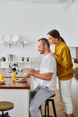 Téléchargez les photos : Vue latérale d'un homme souriant dégustant un délicieux petit déjeuner près d'une femme attentionnée dans la cuisine, couple sans enfant - en image libre de droit