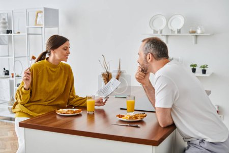 Téléchargez les photos : Femme souriante lisant le journal pendant le petit déjeuner avec mari dans la cuisine moderne, couple sans enfant - en image libre de droit