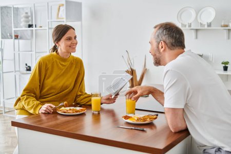 cheerful woman reading newspaper during breakfast with husband in kitchen, child-free lifestyle
