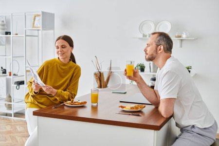 Téléchargez les photos : Femme souriante lire des nouvelles au mari pendant l'heure du petit déjeuner dans la cuisine, matin du couple sans enfant - en image libre de droit