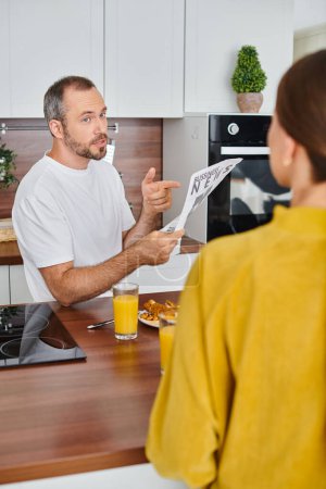 serious man pointing at newspaper near wife during breakfast in kitchen, child-free lifestyle