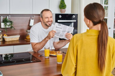 cheerful man showing newspaper to wife during breakfast in modern kitchen, child-free lifestyle