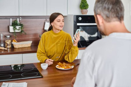 smiling woman reading news from smartphone while having breakfast with husband, child-free couple