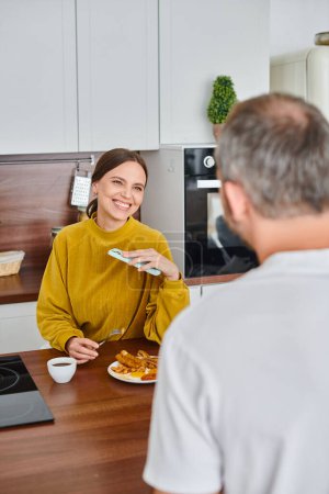 fröhliche Frau mit Smartphone beim Frühstück mit Mann in Küche, kinderfreies Paar