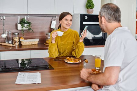 cheerful woman with coffee and smartphone talking to husband during breakfast, child-free lifestyle