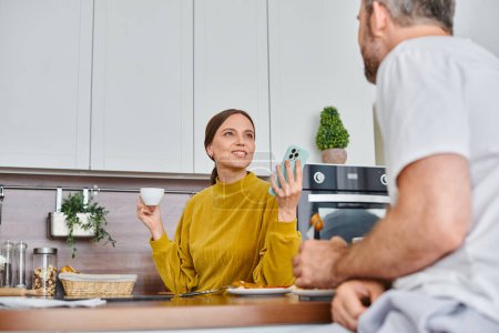 Téléchargez les photos : Femme excitée avec smartphone et tasse de café parler au mari pendant le petit déjeuner dans la cuisine - en image libre de droit