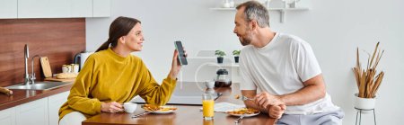 aufgeregte Ehefrau zeigt ihrem Mann während des Frühstücks in der modernen Küche das Smartphone, horizontale Fahne