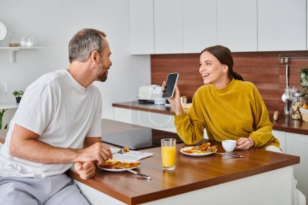 happy woman showing smartphone to husband during breakfast in modern kitchen, child-free couple