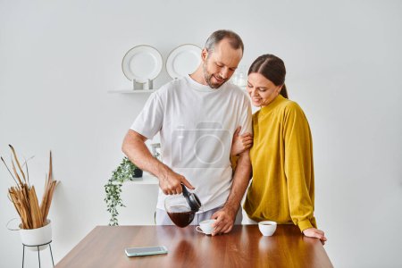 Lächelnder Mann gießt frischen, aromatischen Kaffee in der Nähe seiner Frau in die moderne Küche, am Morgen des kinderfreien Paares
